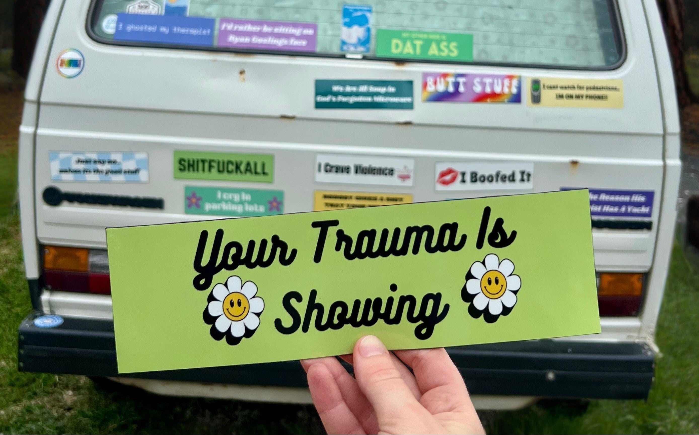 Van with various stickers on the back window, held up a hand with a green sticker reading 'Your Trauma Is Showing'.